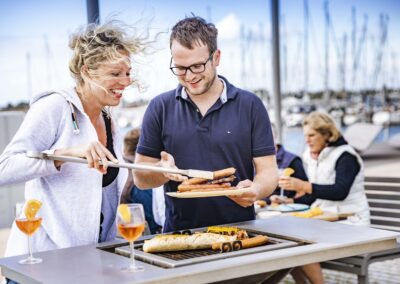 Zwei Personen am Grilltisch mit Würstchen und Baguette vor einem Yachthafen mit Segelbooten im Hintergrund.