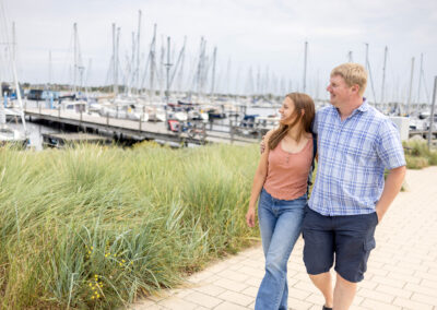 Paar geht Arm in Arm auf gepflastertem Weg entlang eines Yachthafens mit Segelbooten im Hintergrund und Gras am Wegesrand.