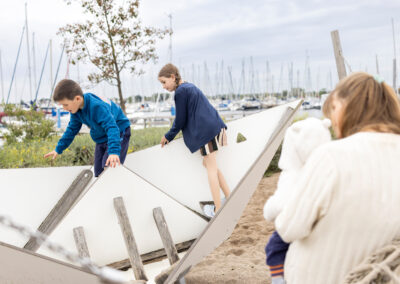 Zwei Kinder klettern auf einem schräg stehenden Spielgerät in Form eines Bootsrumpfs an einem Sandspielplatz am Yachthafen.